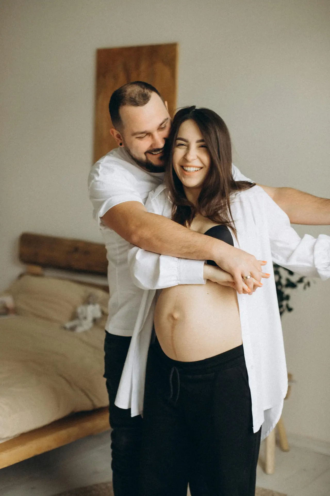 Joyful expectant couple sharing a tender moment in a cozy bedroom setting.
